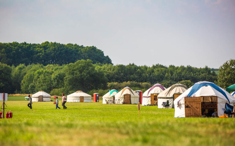 Traditional yurts with different colours are spaced out in Kelmarsh Estate, with trees in the background.