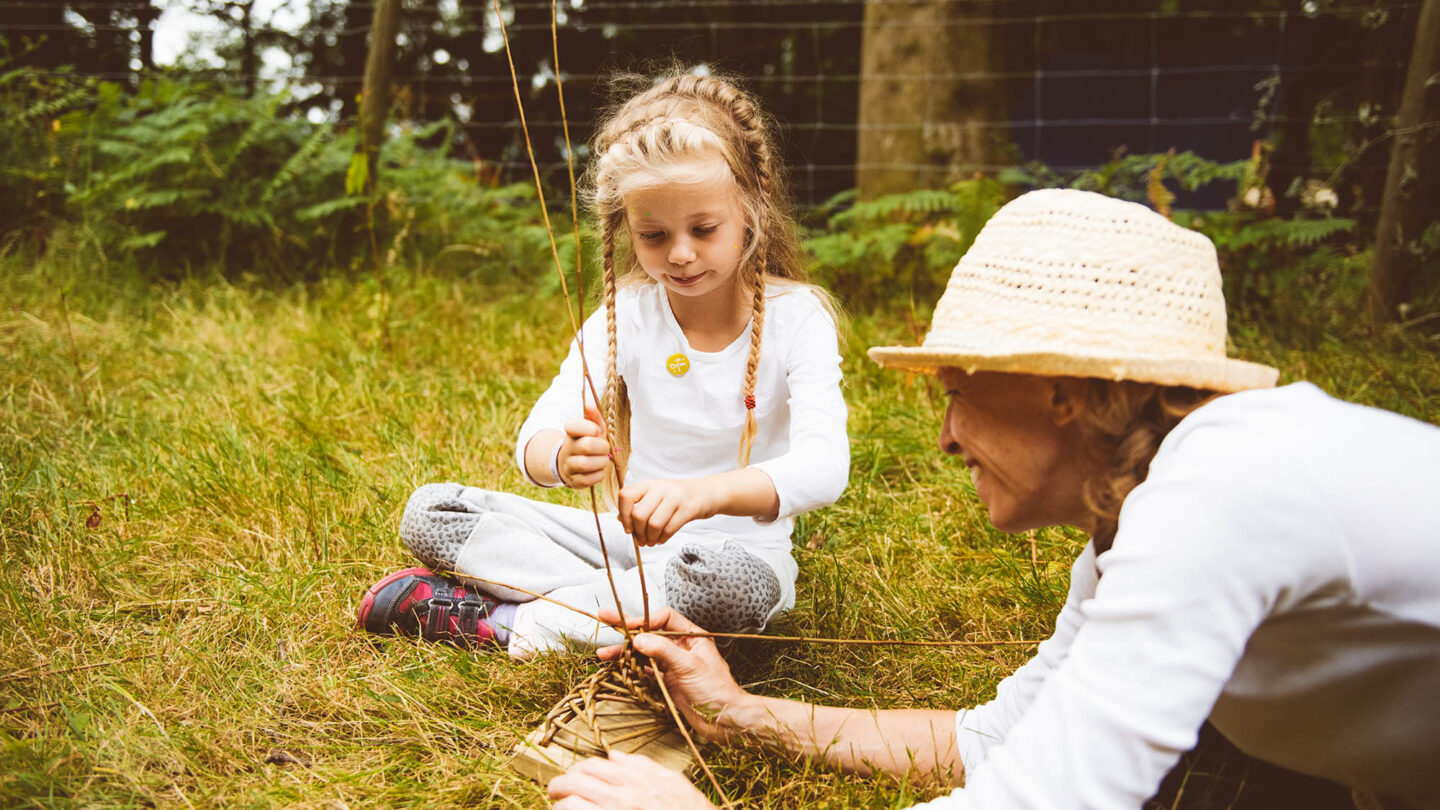 A young girl sits crossed legged twisting willow, while an older women holds the weaved based secure.