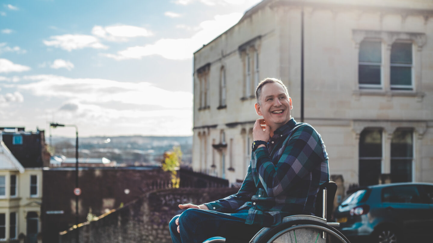 Stephen sits in his wheelchair in front of some buildings. He is wearing a blue checked shirt. He is smiling towards the middle distance