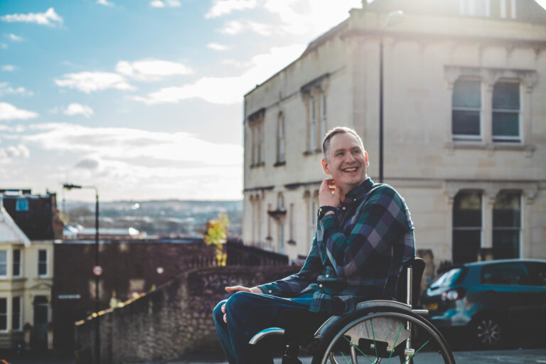 Stephen sits in his wheelchair in front of some buildings. He is wearing a blue checked shirt. He is smiling towards the middle distance